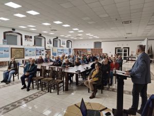 grupa ludzi siedzi na krzesłach przy stole słuchając wykładu